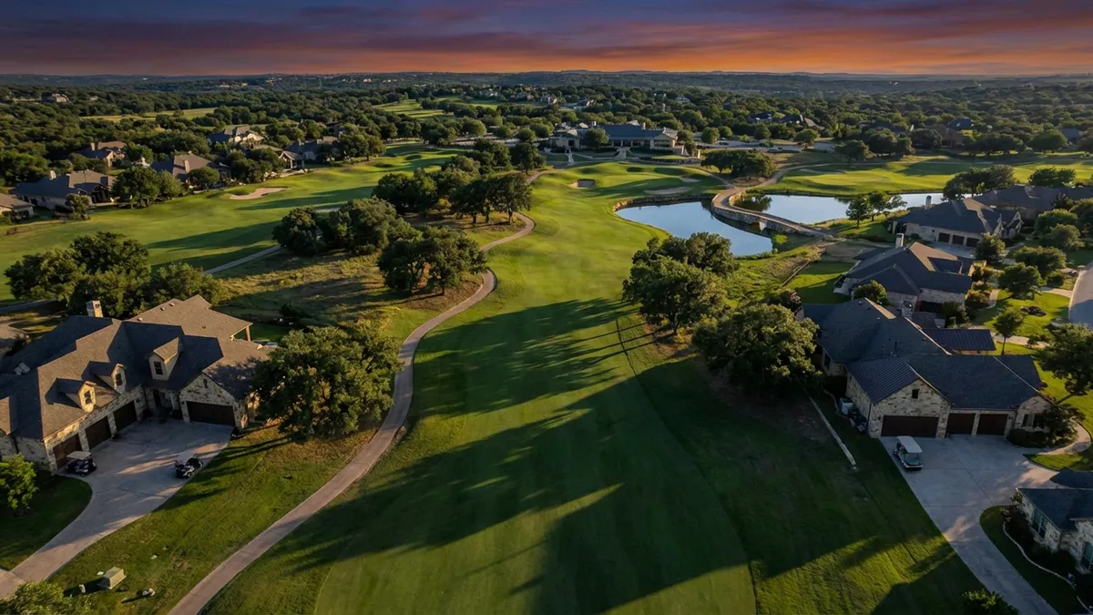 Aerial drone shot capturing the 18th hole at twilight, showcasing emerald fairways cutting through native Texas oaks, with luxury homes featuring stone exteriors and golf cart garages lining the course. Cinematic lighting with long shadows, 16:9 aspect ratio, 8K detail showing individual grass blades and reflective water hazards.