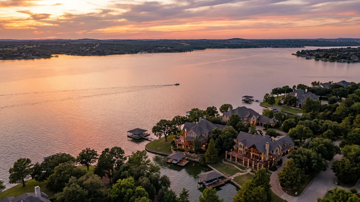 Cinematic aerial view of Lake Lewisville at golden hour, capturing the shimmering water surface reflecting sunset colors. The frame includes Hickory Creek's shoreline with luxury waterfront homes nestled among mature trees, their docks extending into the calm water. In the distance, a fishing boat creates gentle ripples. Ultra HD 8K resolution with rich details of water textures, architectural elements, and natural surroundings. 16:9 aspect ratio, professional real estate photography style with warm, inviting tones.