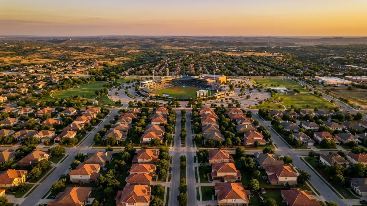 Cinematic aerial view of Round Rock at golden hour, capturing suburban neighborhoods with tree-lined streets, Dell Diamond stadium in midground, and the Texas Hill Country in distance. 16:9 aspect ratio, ultra HD 8k realism with warm sunlight highlighting red tile roofs and lush green spaces. No people visible—focus on clean residential geometry and community infrastructure.