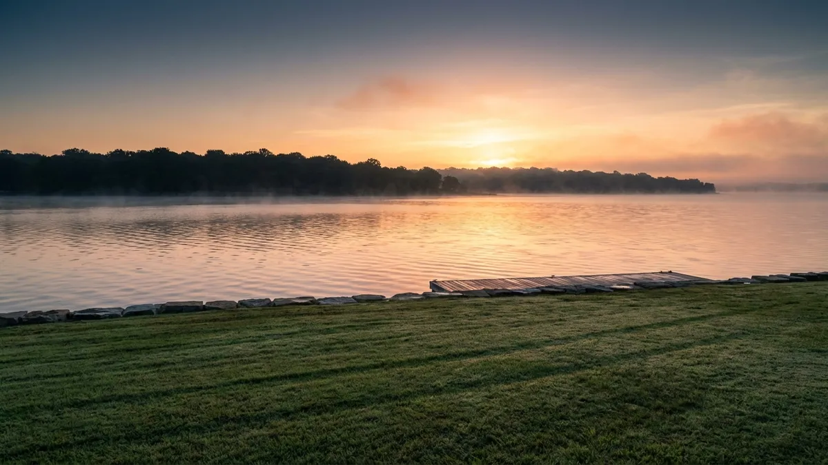 A wide-angle landscape shot capturing the calm waters of Benbrook Lake at dawn. Gentle ripples on the water surface reflect soft, golden-pink hues from the rising sun peeking over a distant treeline. In the foreground, a neatly manicured green lawn with a subtle dew, leading to the lake's edge. The sky transitions from a deep blue at the top to a warm orange-yellow near the horizon. Cinematic lighting, golden hour, 16:9, highly detailed 8k.