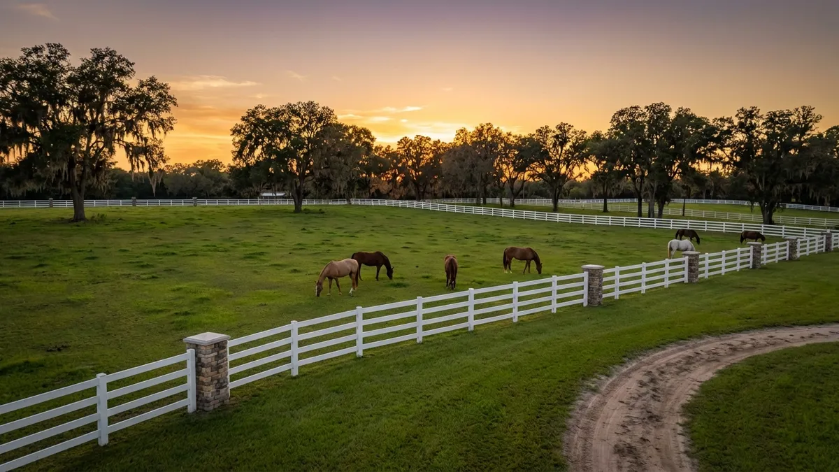 A wide-angle landscape view at golden hour of rolling green pastures with several horses grazing peacefully near a pristine white-fenced ranch. Mature oak trees dot the background, and a glimpse of a well-maintained equestrian trail can be seen winding through the scene. Cinematic lighting, soft shadows, 16:9, highly detailed 8k.