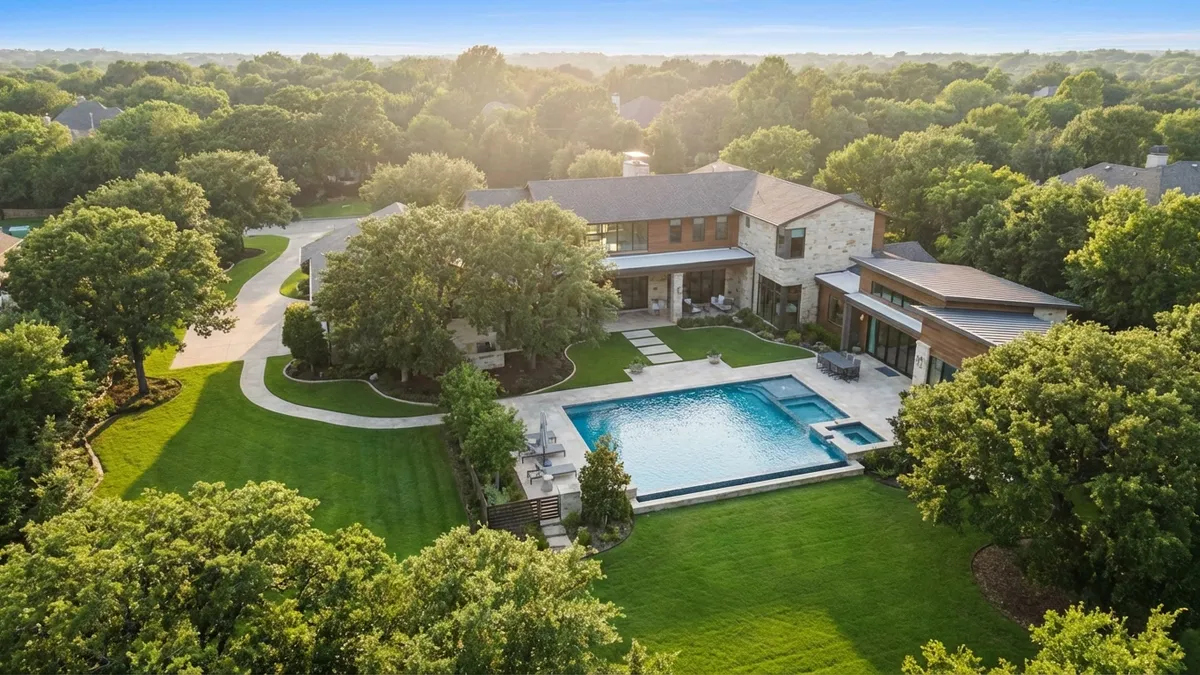Overhead drone shot, modern luxury home in Flower Mound, Texas, surrounded by lush green lawns and mature trees, sparkling swimming pool visible, clear blue sky, soft afternoon sunlight, 16:9 aspect ratio, highly detailed 8k, volumetric lighting, natural colors.