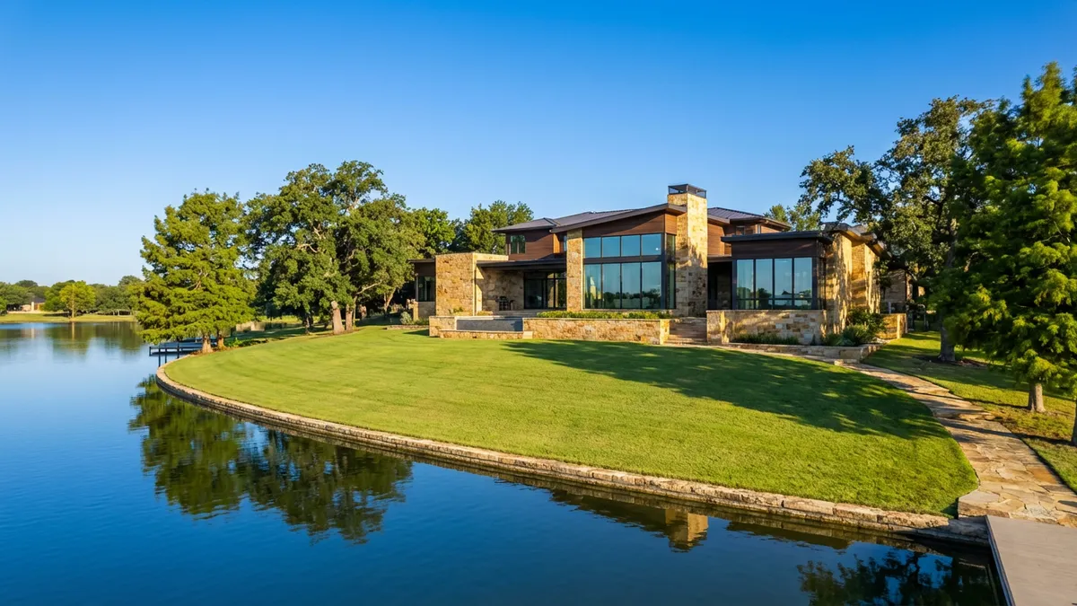A highly detailed wide shot of a modern luxury home overlooking a calm lake, surrounded by lush green trees under a clear blue sky. Cinematic lighting, mid-morning sun, 16:9, highly detailed 8k. The architecture is clean and contemporary, with large windows reflecting the sky and water. Focus on the textures of natural stone and wood. The lake has minimal ripples, mirroring the sky. A pristine, expansive green lawn leads to the water's edge.